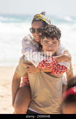 Une femme embrasse un jeune homme avec affection sur une plage de sable, partageant un moment de joie en famille sous le soleil. Banque D'Images