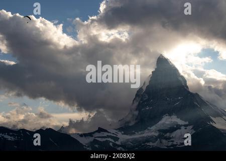 Un parapente vole près du majestueux Cervin, silhouette sur le ciel spectaculaire d'un coucher de soleil suisse, avec des nuages tourbillonnant autour du sommet Banque D'Images