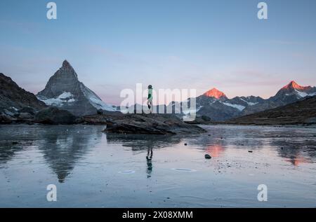 Un individu se tient au bord d'un lac dégelé avec l'emblématique montagne Matterhorn des Alpes suisses illuminé par la lueur de la lumière tôt le matin au printemps. Banque D'Images