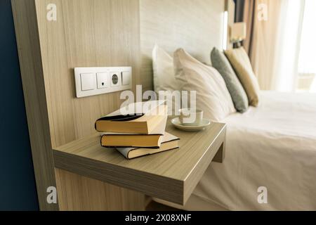 Une pile de livres repose sur une étagère de chevet moderne dans une chambre d'hôtel réconfortante, avec une tasse mise de côté. Banque D'Images