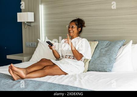 Une joyeuse femme hispanique d'âge moyen dans une robe blanche profite du temps de loisirs sur un lit d'hôtel confortable, en utilisant la télécommande de la télévision avec un sourire ravi. Banque D'Images