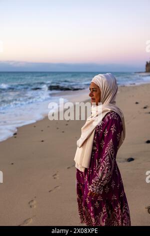 Une femme musulmane paisible dans un hijab se tient sur la plage, regardant l'océan alors que le crépuscule s'installe Banque D'Images