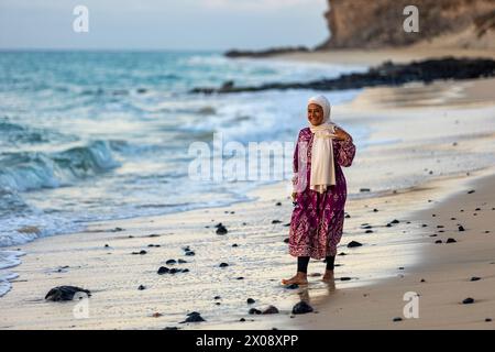 Une femme musulmane joyeuse portant un hijab et une robe traditionnelle marche le long d'une plage de sable, avec des vagues se brisant doucement en arrière-plan Banque D'Images