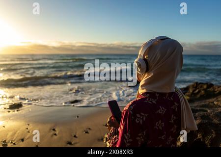 Une femme musulmane en hijab écoute de la musique avec des écouteurs tout en tenant un smartphone, regardant la mer pendant un beau coucher de soleil Banque D'Images