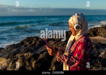 Une femme musulmane souriante portant un hijab aime la musique avec des écouteurs tout en tenant un smartphone sur une côte rocheuse avec des vagues en arrière-plan durin Banque D'Images
