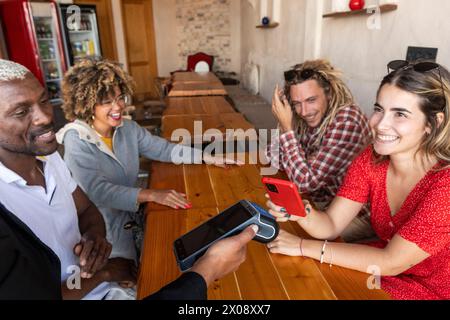 Un groupe joyeux d'amis divers partage un rire autour d'une table en bois dans un cadre de bar confortable, s'engageant avec leurs smartphones Banque D'Images