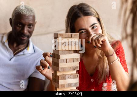 Groupe multiracial d'amis ayant du plaisir à jouer un jeu de bloc en bois à l'intérieur, avec l'accent sur le jeu et le visage souriant d'une femme Banque D'Images