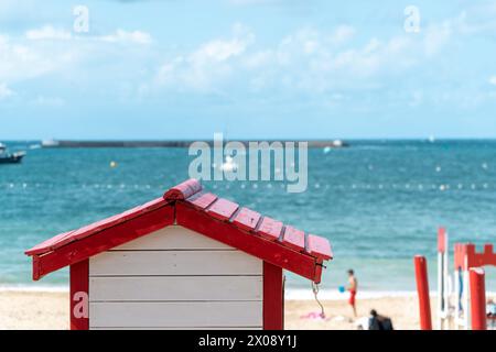 La cabane de plage rouge vif se distingue sur les eaux bleues sereines, avec des bateaux naviguant en arrière-plan à Saint Jean de Luz, en France Banque D'Images
