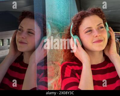 Jeune femme rousse souriante aux cheveux roux bouclés portant une chemise rayée et appréciant la musique avec des écouteurs sarcelle, avec un reflet sur le verre Banque D'Images