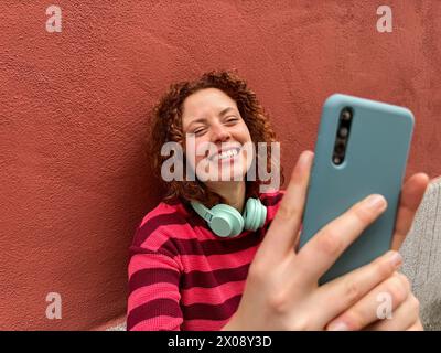 Jeune femme rousse souriante en pull rayé ayant un chat vidéo amusant sur son smartphone sur un fond rouge Banque D'Images
