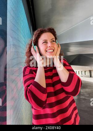Une femme rousse souriante aux cheveux bouclés aime la musique sur son casque, sentant le rythme avec une expression joyeuse sur un fond urbain Banque D'Images