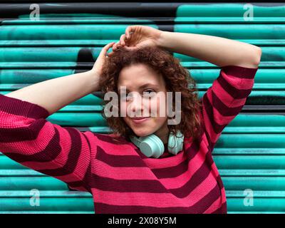 Jeune femme rousse souriante dans un pull rayé posant sur un fond bleu sarcelle vibrant avec des écouteurs autour de son cou Banque D'Images