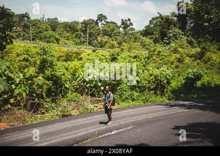 Un voyageur solitaire se tient sur une route désolée au milieu de la végétation luxuriante de la campagne indonésienne, symbolisant l'aventure et l'exploration Banque D'Images