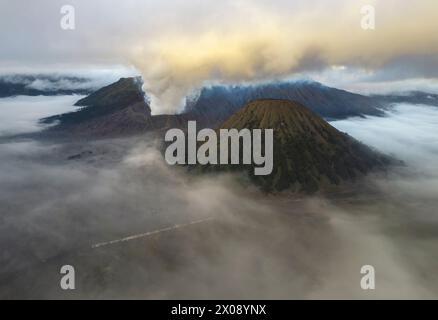 Une vue aérienne éthérée des pics volcaniques émergeant d’une mer de brume à l’aube, avec de la fumée s’élevant d’un cratère actif Banque D'Images