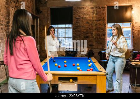 Les jeunes femmes amies profitent d'une partie décontractée de billard dans une salle confortable aux murs de briques Banque D'Images