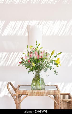 Un bel arrangement de fleurs fraîches dans un vase clair se trouve sur une table en rotin, avec la lumière du soleil projetant des ombres rayées sur un mur blanc Banque D'Images