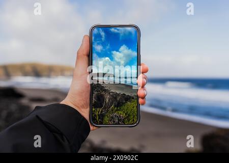 Une main tient un smartphone qui s'aligne parfaitement avec un magnifique paysage de plage, mettant en valeur une perspective créative près de la plantation de thé Gorreana Banque D'Images