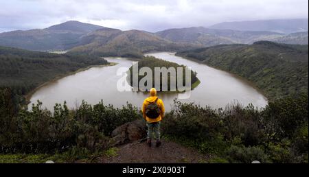 Vue arrière d'un randonneur isolé méconnaissable vêtu d'une veste jaune se dresse sur le bord d'une colline, regardant vers le bas un majestueux méandre de rivière entouré par gre Banque D'Images