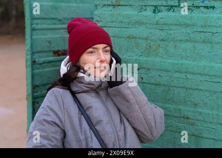 Une jeune femme à la mode vêtue de vêtements d'automne confortables parlant sur un smartphone tout en se tenant à l'intérieur d'une vieille cabine à langer altérée sur un au paisible Banque D'Images