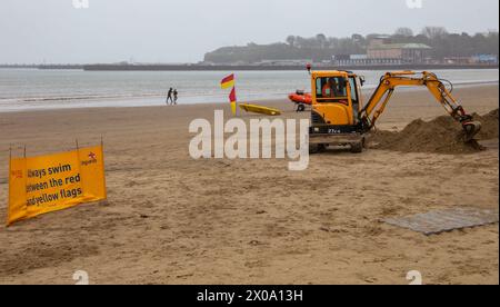 Mini pelle empilant le sable sur la plage à Weymouth Dorset UK par une journée humide et venteuse avec des drapeaux de sauveteur et de sortie. Banque D'Images