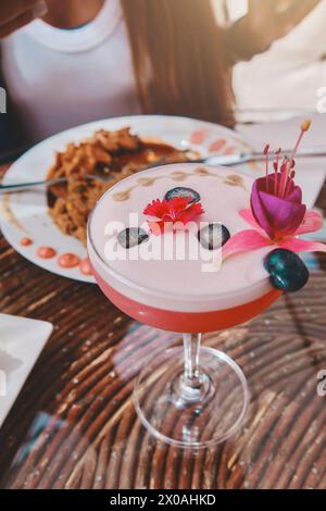 Un verre de boisson rose avec une fleur sur le dessus se trouve sur une table à côté d'une assiette, nourriture et boisson Banque D'Images