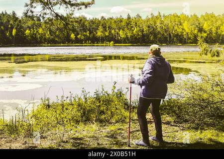 Femme senior stand tenir des bâtons nordiques rouges et profiter de la vue panoramique sur le lac en belle journée ensoleillée. Fond de concept de texture et de bien-être de la nature Banque D'Images