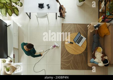 Jeune homme aspirateur plancher quand petite amie regarder un film sur ordinateur portable et manger du pop-corn, vue du haut Banque D'Images