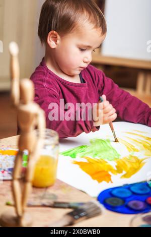 Plan vertical d'un petit enfant peignant avec des peintures acryliques colorées sur sa toile blanche sur sa table de travail, où il y a différents pinceaux, peintures A. Banque D'Images