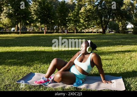 Une femme afro-américaine sinueuse en vêtements de sport s'assoit paisiblement sur une serviette dans un parc, prenant un moment pour se reposer et admirer la nature. Banque D'Images