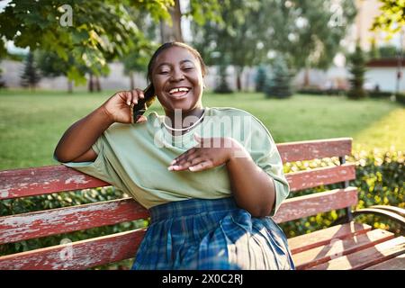 Femme afro-américaine de taille plus en tenue décontractée, assise sur un banc à l'extérieur en été, parlant sur un téléphone portable. Banque D'Images