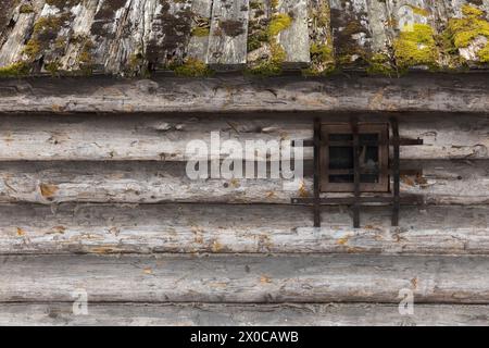 Vieux toit de maison en bois avec mousse verte et mur en rondins gris rugueux avec petite fenêtre, texture de photo de fond, vue de face Banque D'Images