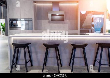 Cuisine dans une nouvelle maison de luxe avec un îlot blanc avec un évier en acier et plaque à induction, à côté de chaises noires et tabourets. Banque D'Images