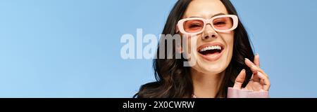 Une jolie femme portant des lunettes de soleil roses dans un studio sur fond bleu, faisant une expression comique. Banque D'Images