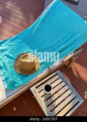 Chaise longue en bois avec serviette bleue, chapeau de paille et tasse de café sur une petite table Banque D'Images