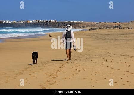Homme solitaire et son chien marchant le long d'une plage de sable laissant des empreintes, El Cotillo, Fuerteventura. Prise en février 2024 Banque D'Images