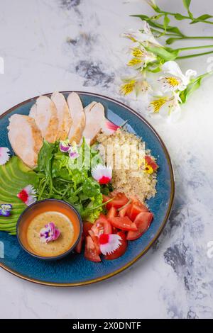 Vue de dessus du déjeuner nutritif avec poulet grillé, salade, tomates, avocat, sauce couscous sur plaque bleue. Banque D'Images