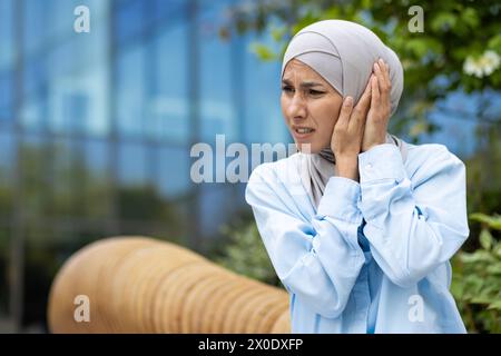 Femme d'âge moyen dans un hijab bleu clair tenant son oreille dans la douleur à l'extérieur, avec une expression préoccupée, peut-être souffrant d'otite ou d'inflammation de l'oreille. Banque D'Images