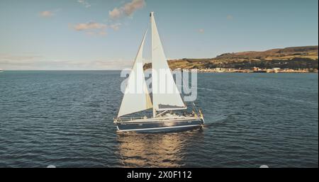 Voile au yacht de course en pleine mer, soleil brillant vue aérienne. Paysage marin majestueux sur la côte de l'île d'Arran avec fond de ciel bleu. île vallonnée d'Écosse avec jetée Brodick. Scène cinématographique luxueuse Banque D'Images