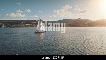 Voile au yacht de course en pleine mer, soleil brillant vue aérienne. Paysage marin majestueux sur la côte de l'île d'Arran avec fond de ciel bleu. île vallonnée d'Écosse avec jetée Brodick. Scène cinématographique luxueuse Banque D'Images