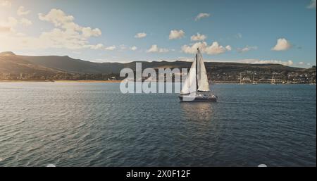 Voile au yacht de course en pleine mer, soleil brillant vue aérienne. Paysage marin majestueux sur la côte de l'île d'Arran avec fond de ciel bleu. île vallonnée d'Écosse avec jetée Brodick. Scène cinématographique luxueuse Banque D'Images