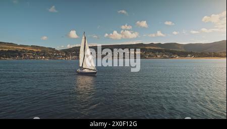 Voile au yacht de course en pleine mer, soleil brillant vue aérienne. Paysage marin majestueux sur la côte de l'île d'Arran avec fond de ciel bleu. île vallonnée d'Écosse avec jetée Brodick. Scène cinématographique luxueuse Banque D'Images