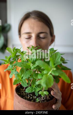 Femme avec les yeux fermés appréciant sentir la plante de menthe verte cultivant à la maison. Passe-temps vert Banque D'Images