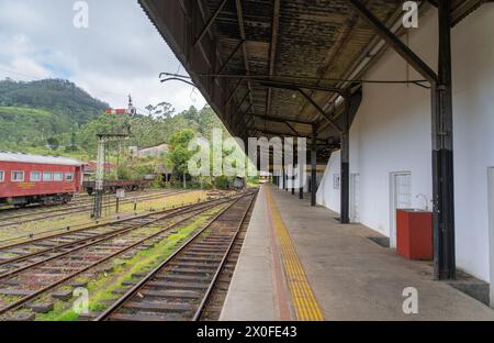 Une gare tranquille, avec des pistes vides menant l'œil vers des montagnes verdoyantes. Un train rouge se repose, évoquant un sentiment de calme. Ambewela, Banque D'Images