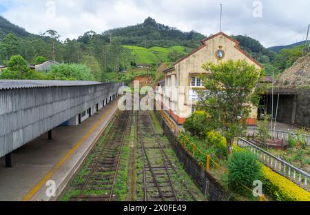 Une gare tranquille, avec des pistes vides menant l'œil vers des montagnes verdoyantes. Un train rouge se repose, évoquant un sentiment de calme. Ambewela, Banque D'Images