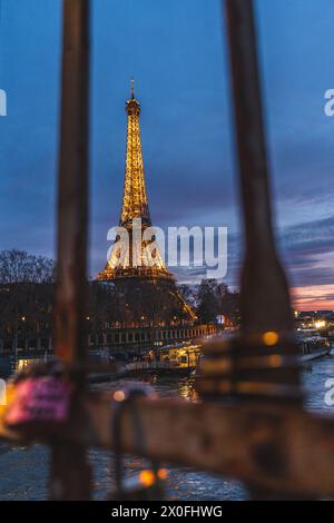 La Tour Eiffel vue entre les barres métalliques d'une clôture remplie de serrures d'amour brille contre le ciel nocturne, projetant une lueur chaude sur la ville parisienne. Banque D'Images