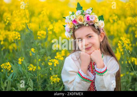 Rêve belle jeune fille avec un chapelet de fleurs, robe folklorique ethnique avec broderie bulgare traditionnelle pendant le coucher du soleil sur un rapseed agricole Banque D'Images