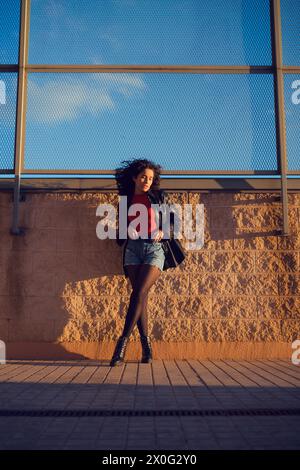 Une femme aux cheveux bouclés pose devant un mur au coucher du soleil Banque D'Images