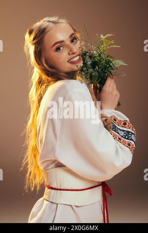 Une jeune fille dans une robe blanche tient délicatement une fleur gracieuse dans un cadre de studio serein. Banque D'Images