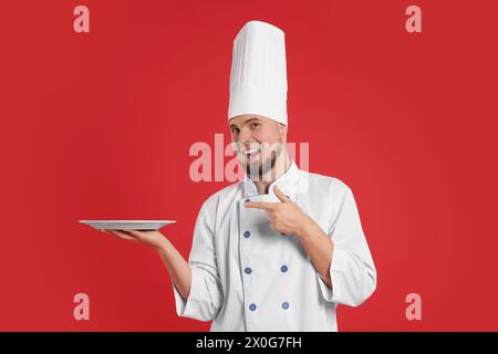 Heureux confiseur professionnel en uniforme pointant à la plaque vide sur fond rouge Banque D'Images