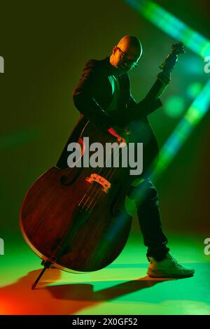 Musicien chauve jouant la contrebasse en néon rouge-vert, lumière de scène sur fond de studio dégradé. Banque D'Images